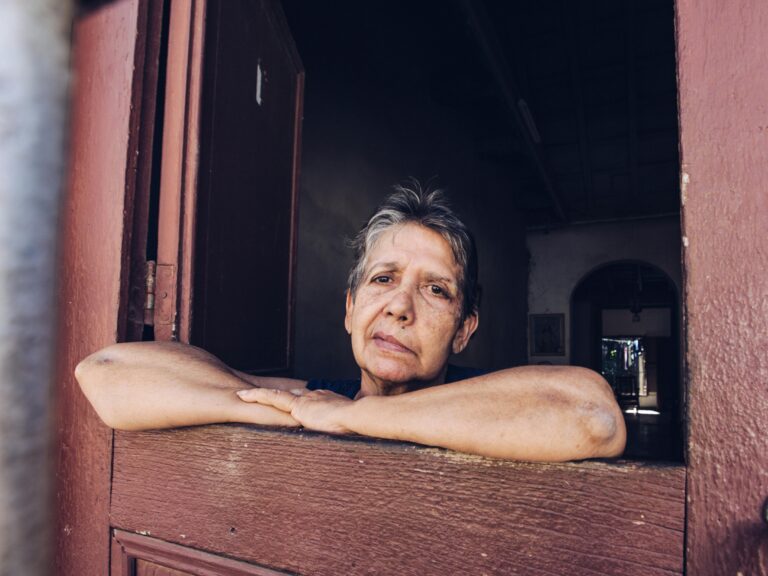 Baby Boomer Woman lookes out a wooden frame window with worried expression on her face.