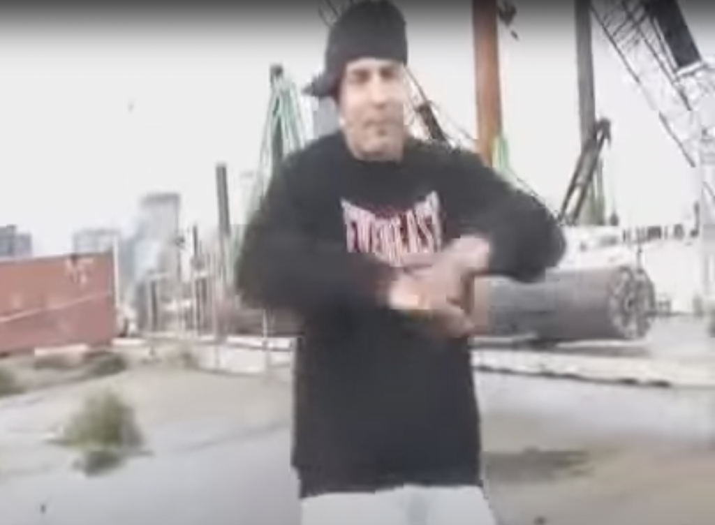 screenshot of man from rap video with backwards black cap in an industrial setting