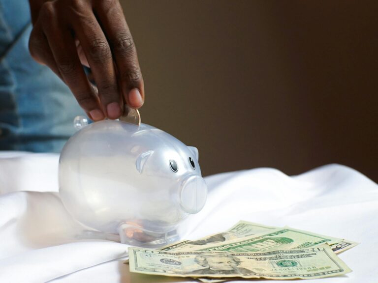 hand putting change in a piggy bank on a table with cash on laying on it.