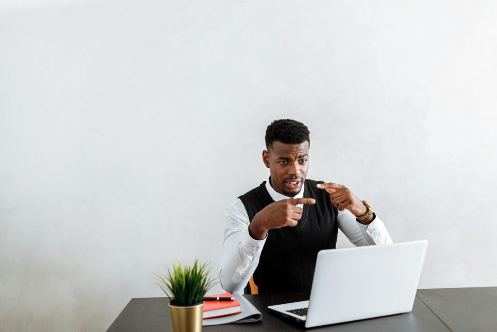 Confused looking professional behind a desk with a laptop and gesturing.