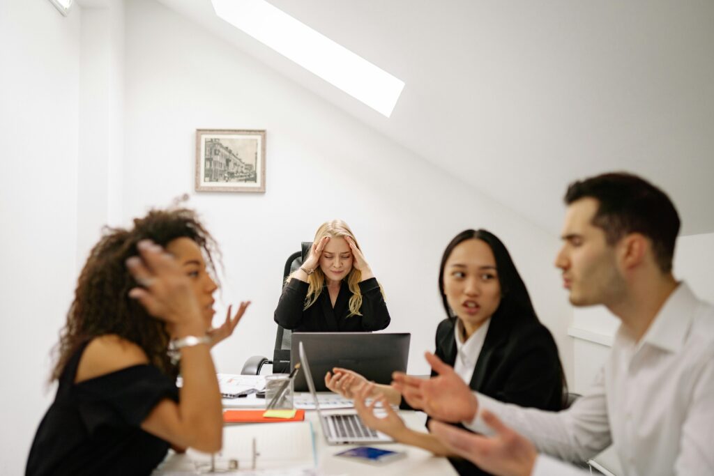 two coworkers arguing at a table while other coworkers watch, looking distressed.