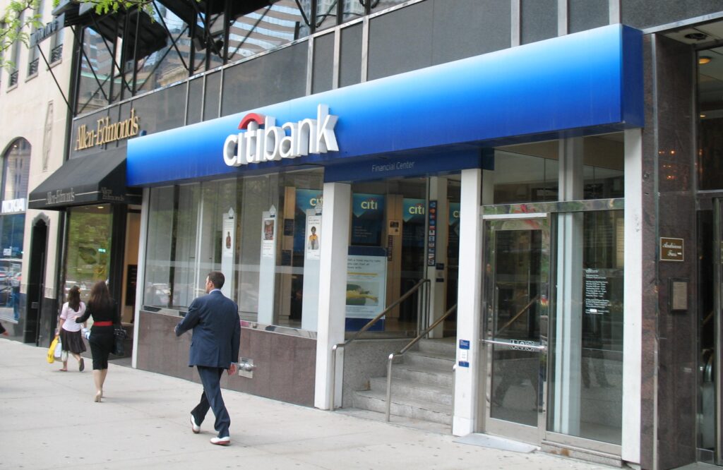 man on city sidewalk walks in front of a citibank storefront