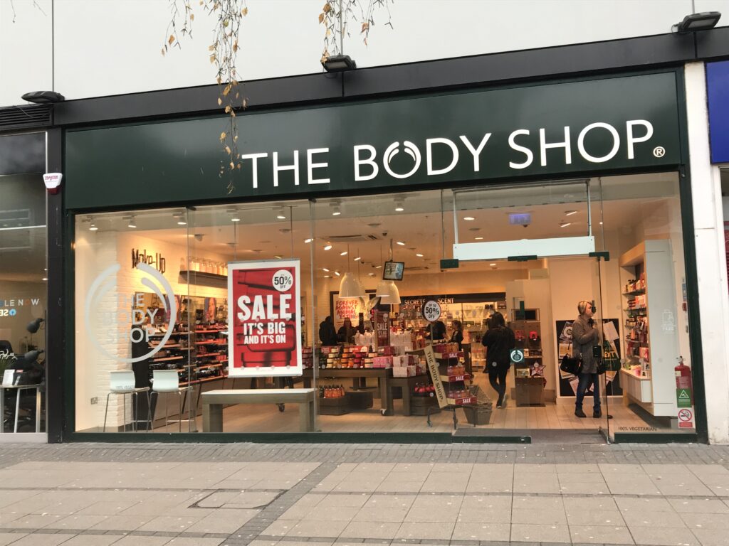 Store front of a The Body Shop Location in West Orchards, Coventry with a Sale sign in the window and lights on. 