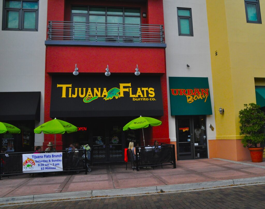 Store front of a Tijuana Flats Mexican restaurant with tables and green umbrellas out front. 