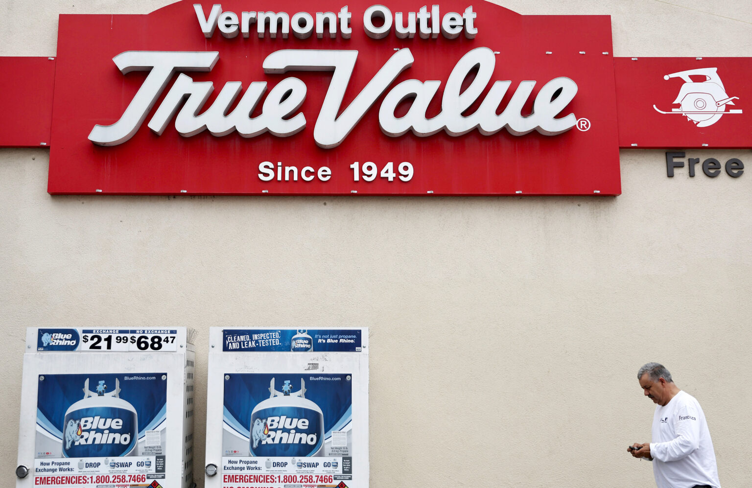 man looking at his cell phone walks in front of a True Value home improvement store. Propane tanks on the left-hand side of the image.