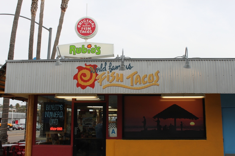 Outside, front-facing photo of the first location of Rubio's Mexican Grill store front against a grey sky in San Diego, California