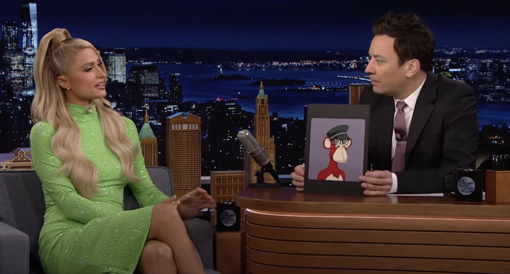 Paris Hilton in a green dres sitting across from Fimmy Fallon in a dark suit behind a desk holding a framed illustration of a monkey NFT on The Tonight how