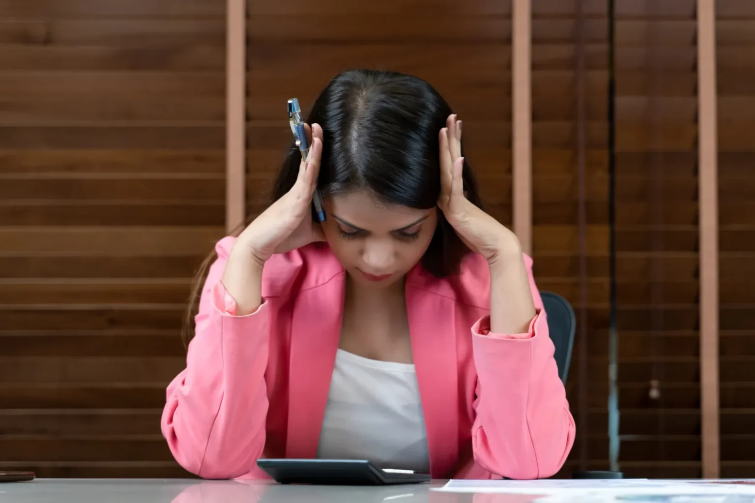 Woman in a link blazer sits at a desk with her head in her hands and looks stressed.