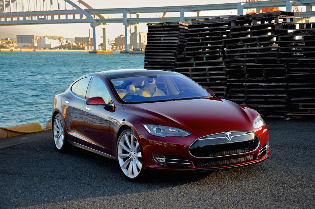 A red Tesla S model parked outside in front of a waterway and bridge