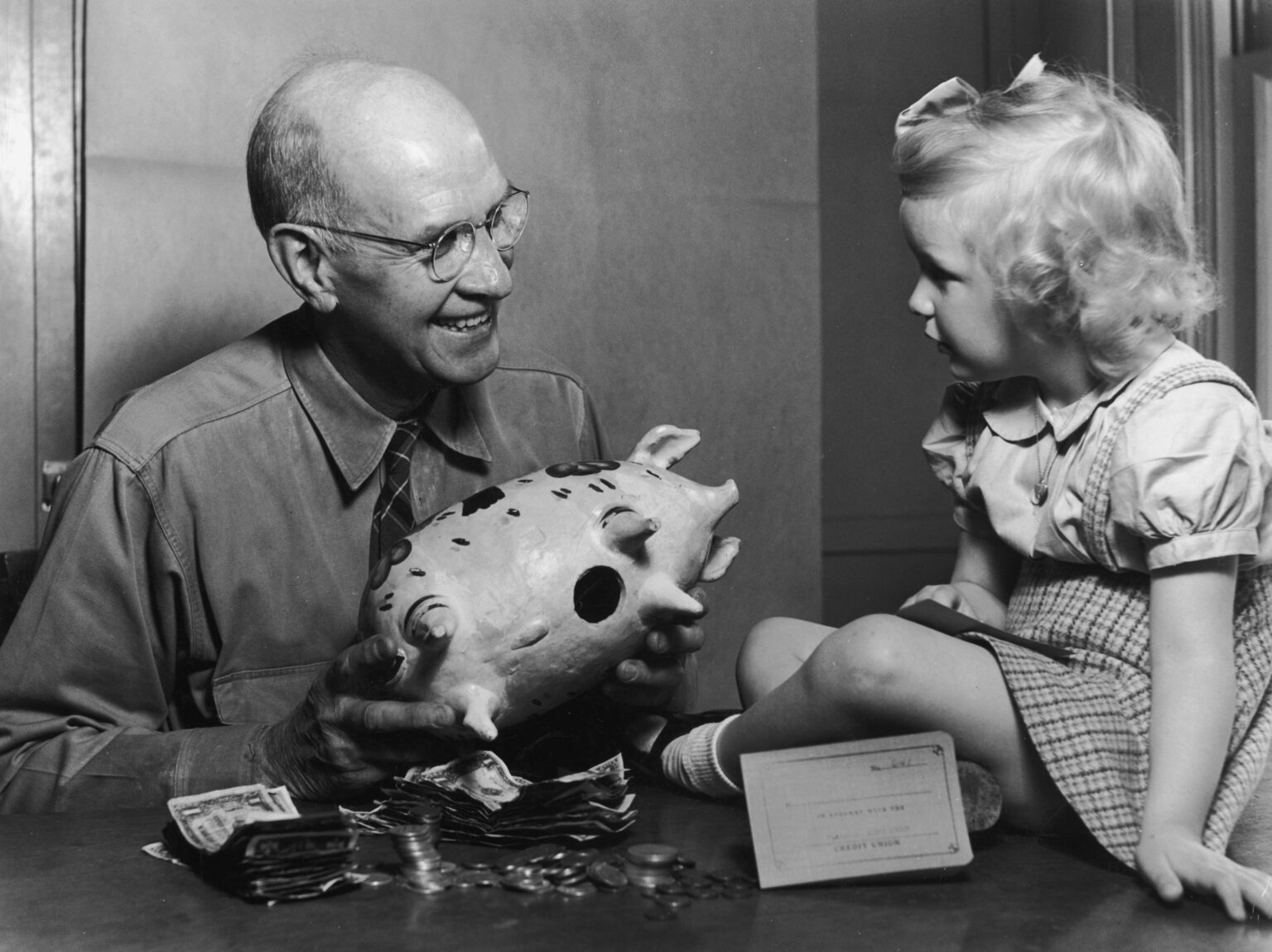 circa 1955: A grandfather smiles as he holds a ceramic piggy bank, helping to unload money for his young granddaughter, who sits and looks on, 1950s.